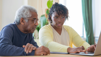 An older adult and a caregiver sit together at a table, reviewing information on a laptop in a bright, plant-filled room.