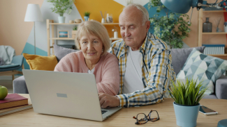 An older couple sits together at a table, using a laptop in a comfortable living room with plants, books, and cozy furnishings.