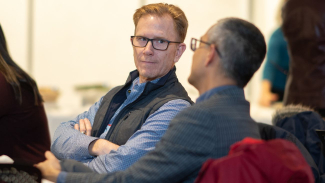 Two people seated indoors at a table during an event or meeting, with one person in the foreground wearing a patterned shirt and vest, and another person turned slightly to the side in conversation.