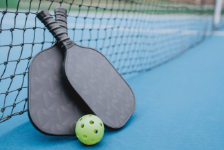 Two pickleball paddles leaning against a net on an outdoor court with a green pickleball in front of them.