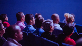 Audience seated in a darkened auditorium listening to a presentation.