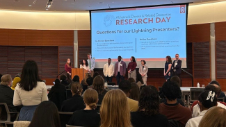 Panel of presenters standing on a stage in front of a projected slide reading ‘Alzheimer’s Disease & Related Dementias Research Day – Questions for our Lightning Presenters?’ as an audience listens in a lecture hall.
