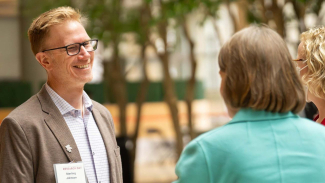 Researcher wearing a “Research Day” name badge that reads “Sterling Johnson” speaks with two colleagues in an indoor atrium with trees in the background.