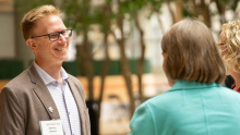 Researcher wearing a “Research Day” name badge that reads “Sterling Johnson” speaks with two colleagues in an indoor atrium with trees in the background.