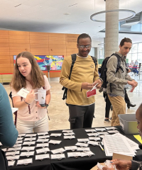 Attendees check in at a conference registration table covered with name badges inside a bright lobby, with several people wearing backpacks and holding programs while staff distribute badges.